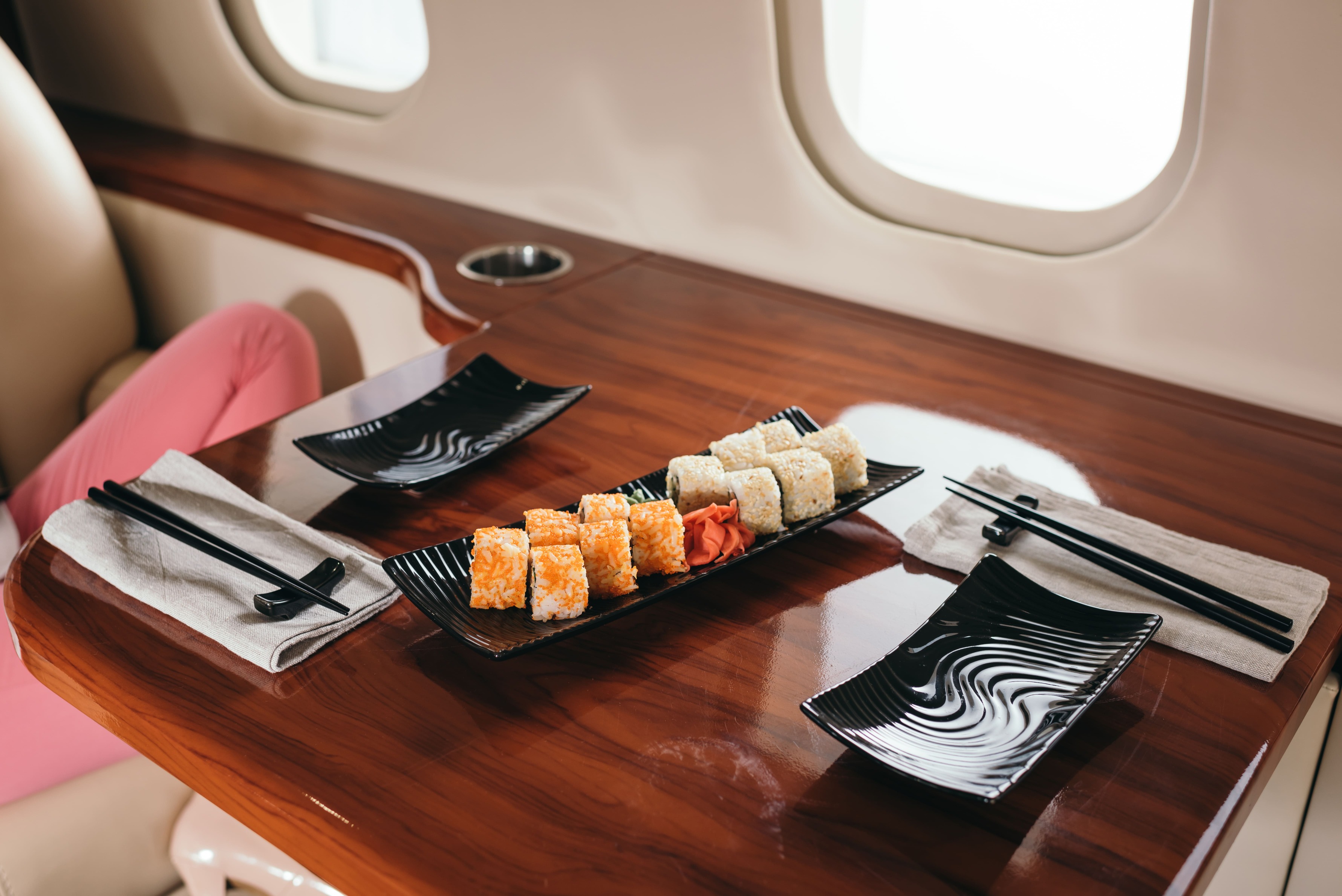 A woman sits in an airplane, enjoying sushi with chopsticks, surrounded by airplane interior elements.