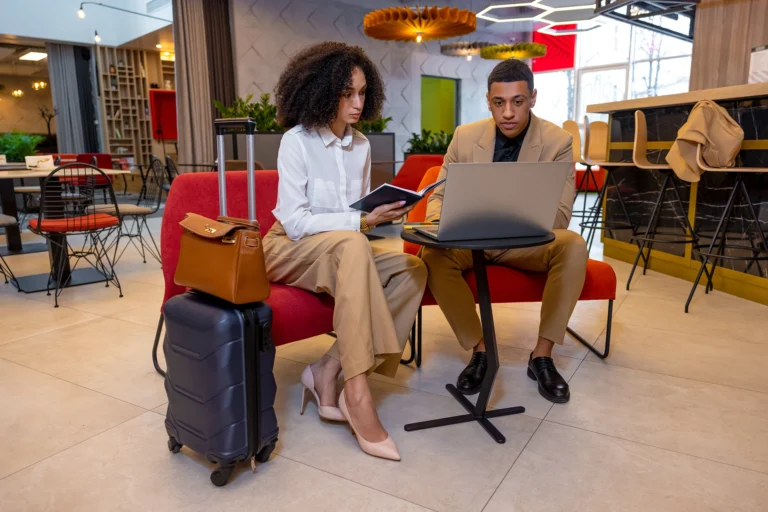 Two business travelers sit on red lounge chairs in a modern hotel lobby, reviewing notes and a laptop beside a rolling suitcase and handbag.