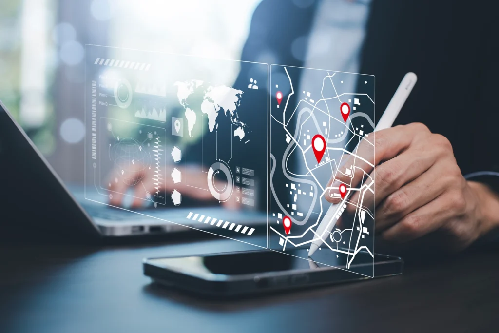 A person using a white stylus on a smartphone to interact with a glowing, holographic interface. The display features a digital world map, data charts, and a street map with red location pins, set against a blurred office background with a laptop.