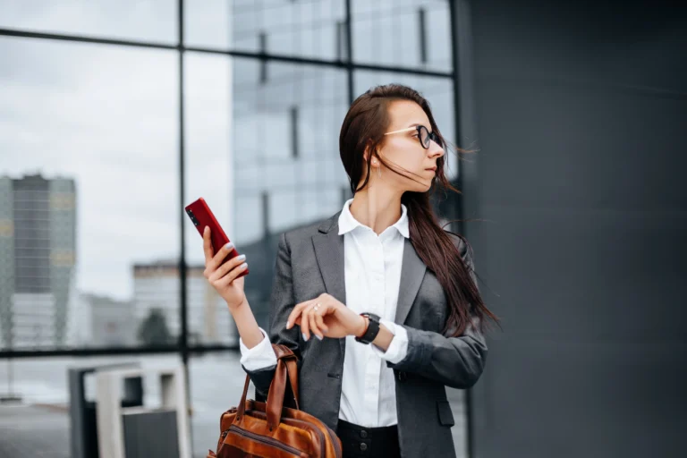 Woman in a gray blazer holding a smartphone and leather handbag, standing outside a modern glass office building and looking to the side.