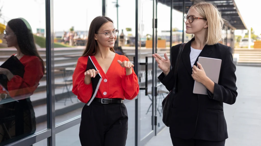 Two women stand outside a modern building with large glass windows, engaged in a conversation while walking.