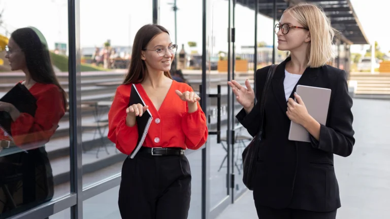 Two women stand outside a modern building with large glass windows, engaged in a conversation while walking.