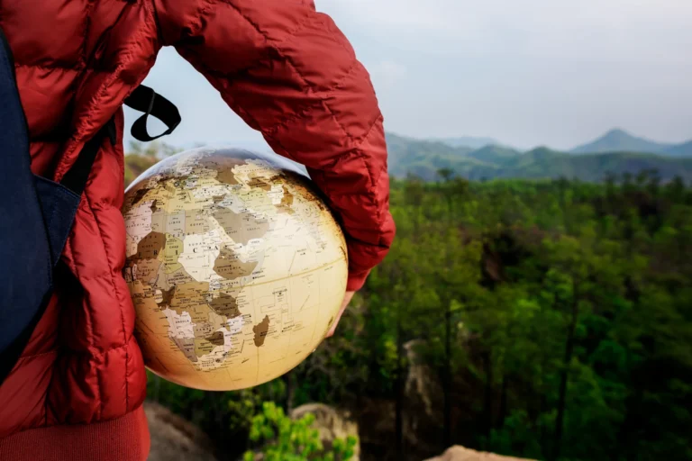Person in a red jacket holding a globe outdoors, with green forest and distant mountains in the background.