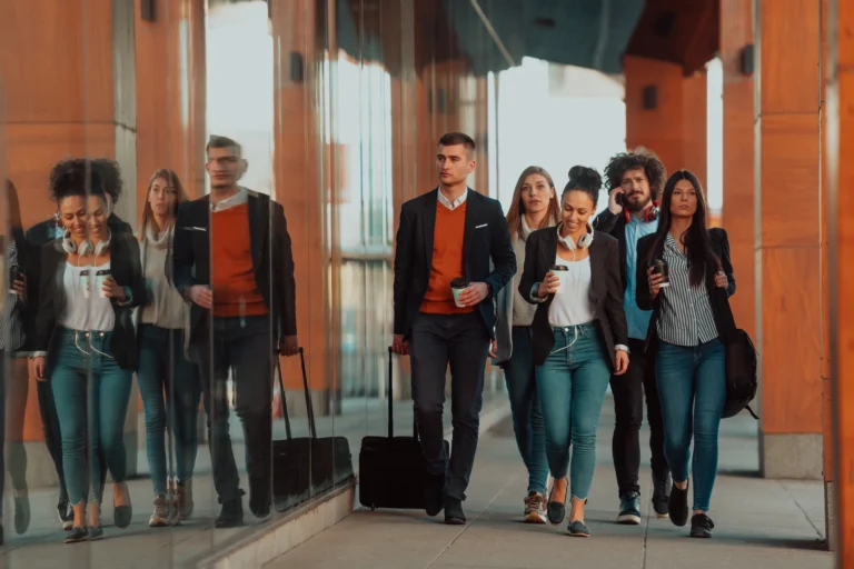 A group of six young professionals walking together outside a modern office building, some holding coffee cups and luggage, with glass walls reflecting their movement.