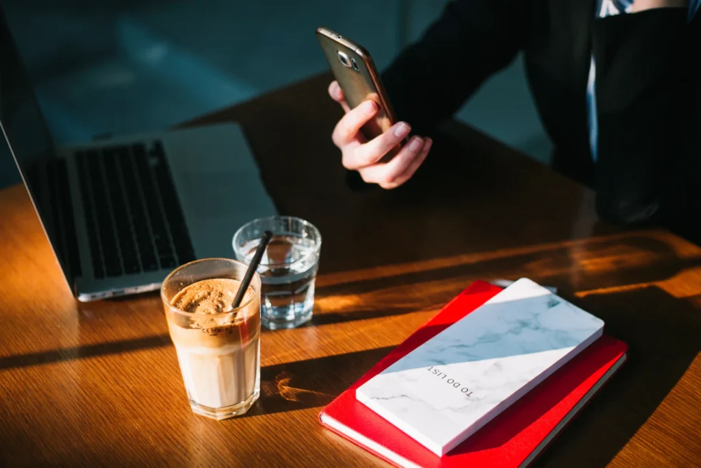 A person in a business suit holding a gold smartphone over a wooden desk bathed in natural afternoon sunlight. The workspace is organized with a laptop, a glass of water, and a frothy iced coffee. In the foreground, a red notebook sits beneath a white marble-patterned 