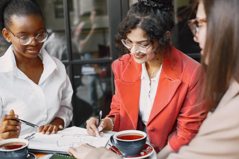 Three women in a meeting reviewing financial charts and documents over coffee, collaborating and discussing data in a modern office setting.