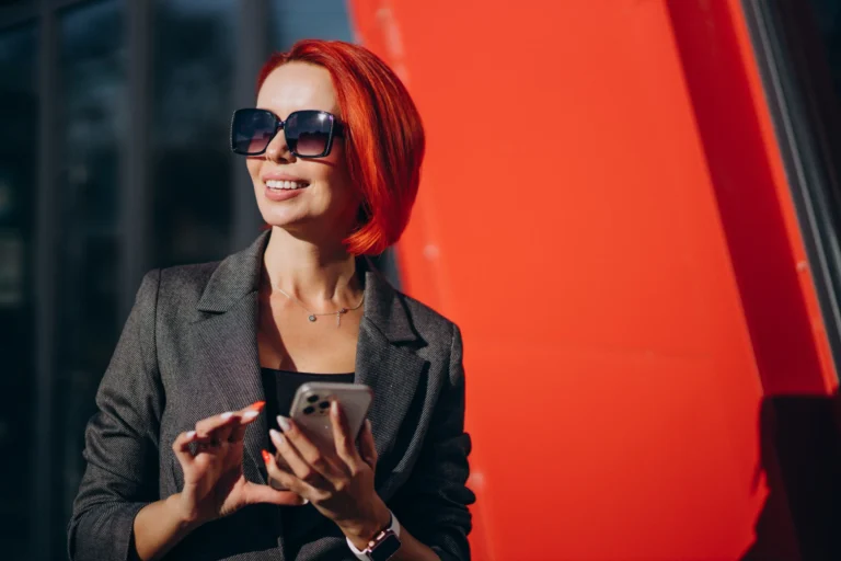 A stylish woman with vibrant red hair and oversized sunglasses smiling while using her smartphone outdoors. She is dressed in a grey blazer and stands against a bold, modern red architectural backdrop. The bright sunlight and her confident expression capture a sense of seamless, tech-enabled travel and modern connectivity.