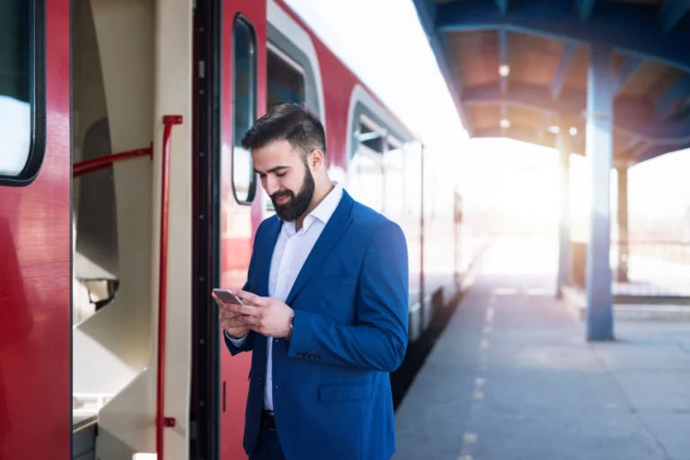 A bearded businessman in a sharp blue suit standing on a sunlit train platform. He is smiling while looking at his smartphone, presumably checking travel details or carbon tracking data. A modern red and white train is stopped behind him, symbolizing sustainable business travel and eco-friendly transportation.