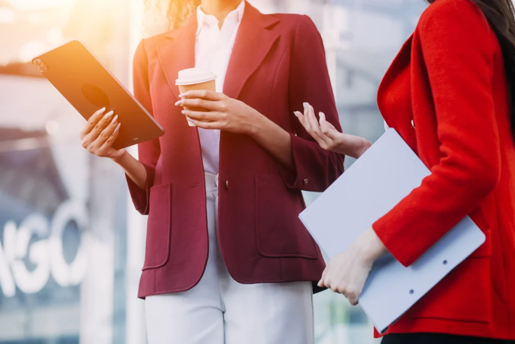 Two businesswomen in red blazers standing outdoors, one holding a tablet and coffee cup while the other holds a folder, engaged in conversation.