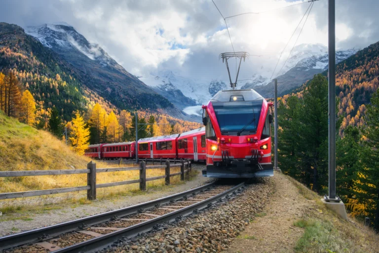 Red passenger train on a curved railway track through autumnal mountains with a wooden fence in the foreground and snow-capped peaks in the distance, under a cloudy sky