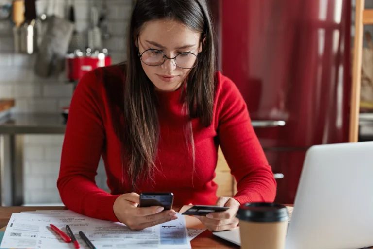 A focused young woman with round glasses and a bright red sweater sitting at a wooden table, managing her business expenses. She is holding a credit card in one hand and typing on her smartphone with the other. Her workspace is cluttered with paper invoices, pens, an open laptop, and a takeaway coffee cup, capturing a relatable moment of remote work or tax preparation in a modern kitchen setting.